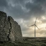 Modern wind turbine spinning strongly in powerful wind while a cracked stone wall stands rigid under dark storm clouds, symbolising adaptability versus resistance in business strategy.